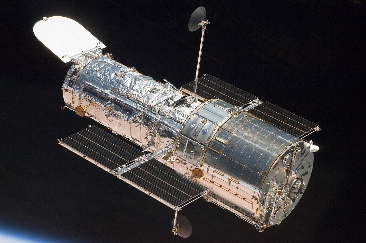The silver cylindrical Hubble Space Telescope with its aperture door open and solar panels extended, against black space.