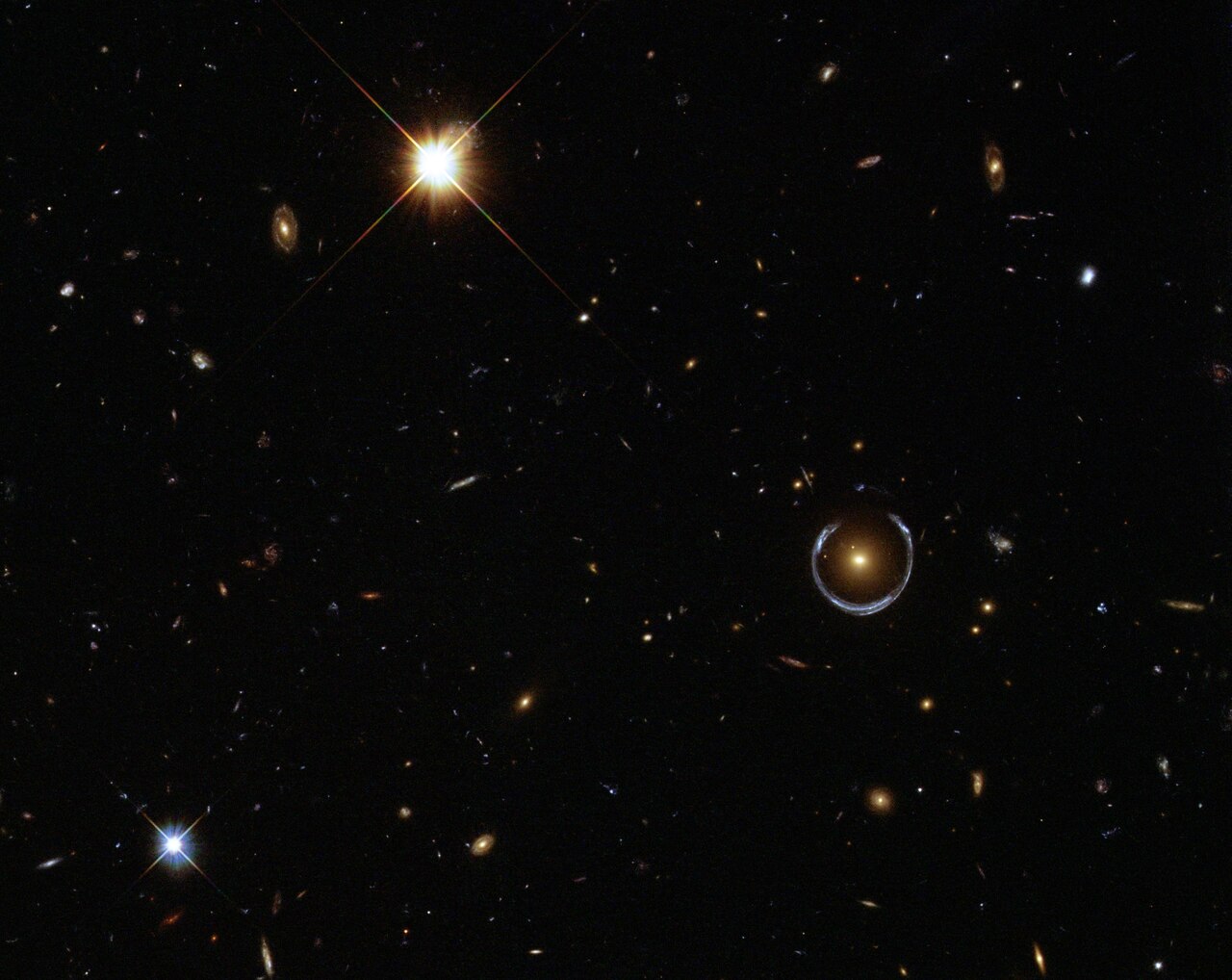 A pale blue, nearly circular arc surrounds a reddish galaxy right of centre, in a dark field scattered with galaxies and bright stars.
