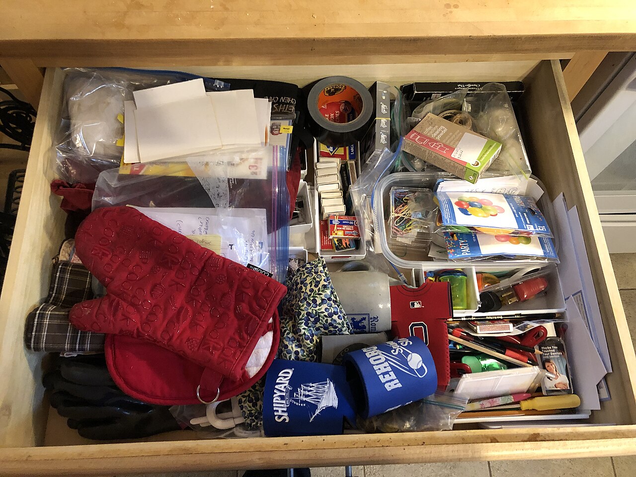 A kitchen junk drawer filled with stationery, small tools, oven mitts and assorted household objects.